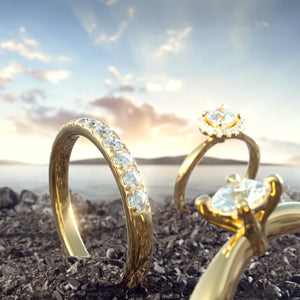 Bild på tre stycken förlovningsringar i guld med labbodlade diamanter som är nedstuckna i sanden. En vacker kvällshimmel i bakgrunden. 