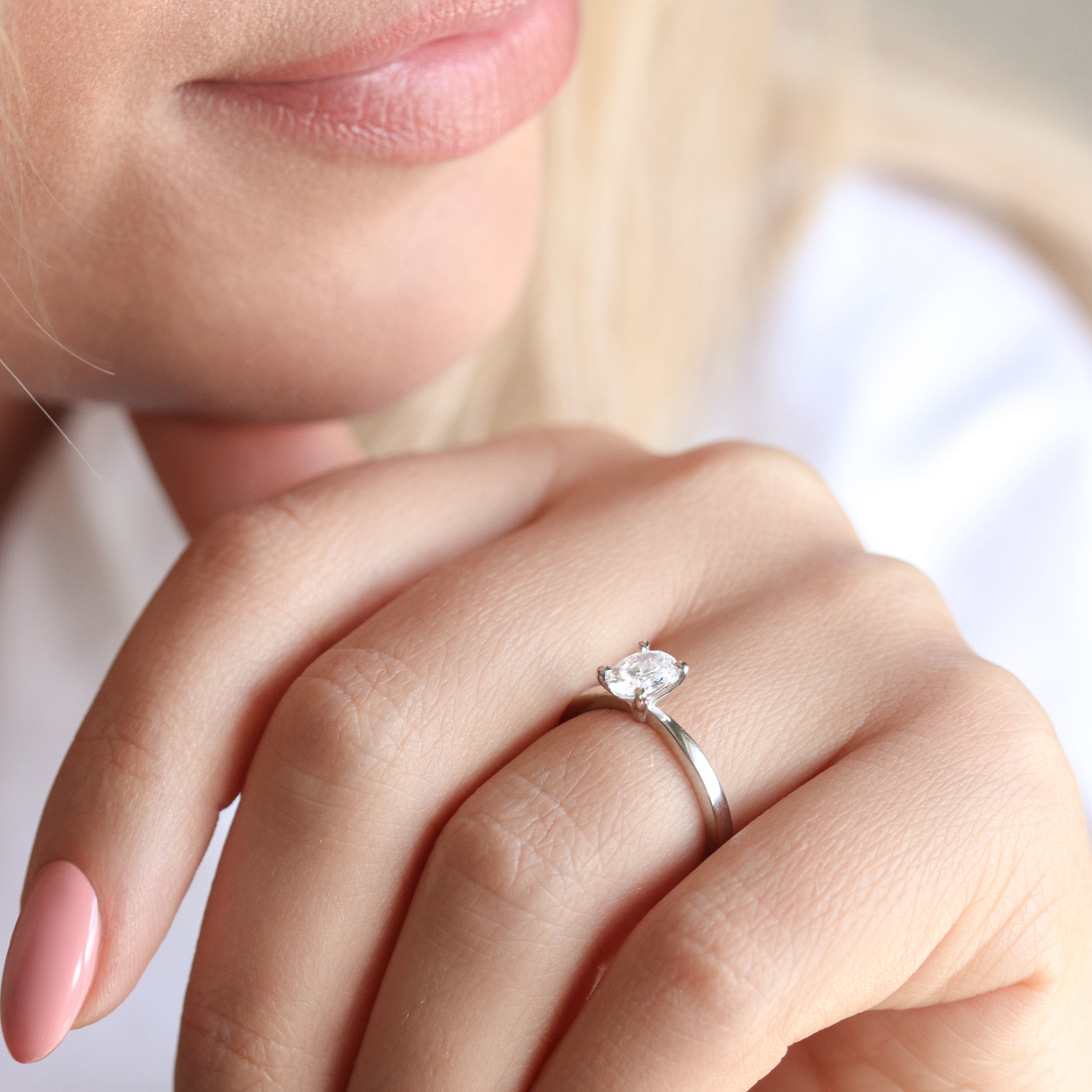 Närbild på en hand som bär en elegant förlovningsring med en oval labbodlad diamant (lab grown diamond) i solitärfattning på ett smalt band av vitguld eller platina. Ringen sitter på ringfingret och visas mot en mjuk, ljus bakgrund som framhäver diamantens briljans.