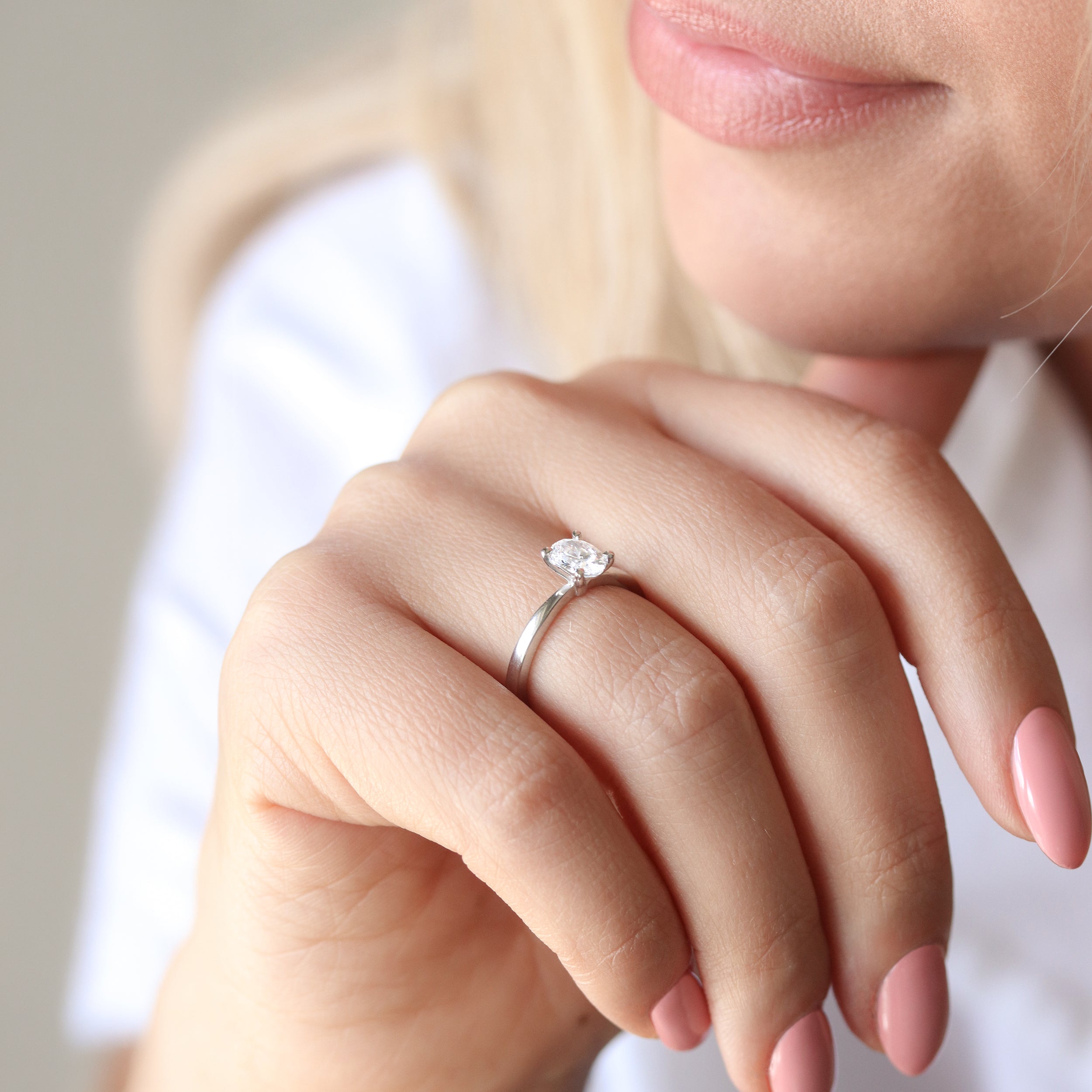 Närbild på en hand som bär en elegant förlovningsring med en oval labbodlad diamant (lab grown diamond) i solitärfattning på ett smalt band av vitguld eller platina. Ringen sitter på ringfingret och visas mot en mjuk, ljus bakgrund som framhäver diamantens briljans.