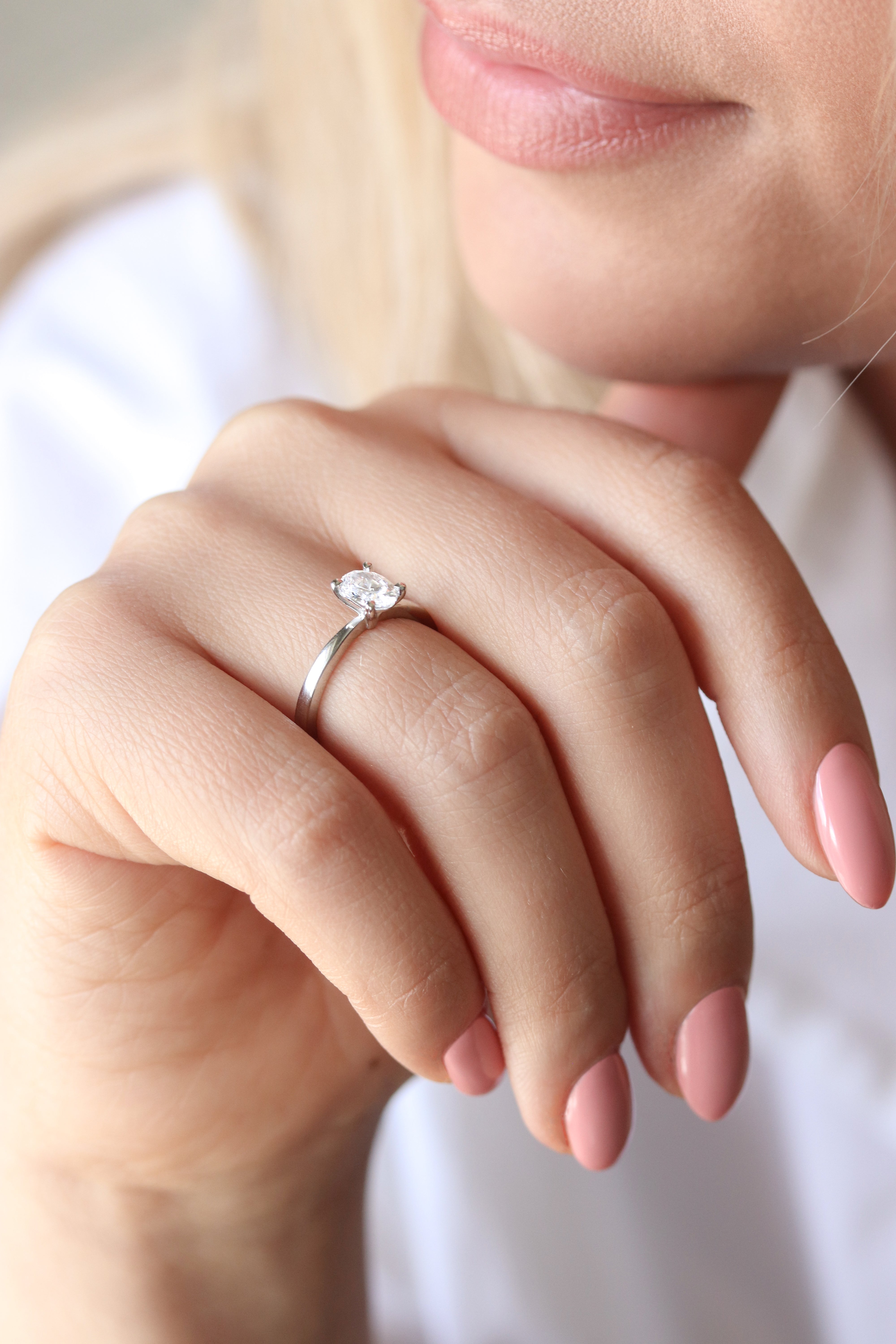 Närbild på en hand som bär en elegant förlovningsring med en oval labbodlad diamant (lab grown diamond) i solitärfattning på ett smalt band av vitguld eller platina. Ringen sitter på ringfingret och visas mot en mjuk, ljus bakgrund som framhäver diamantens briljans.