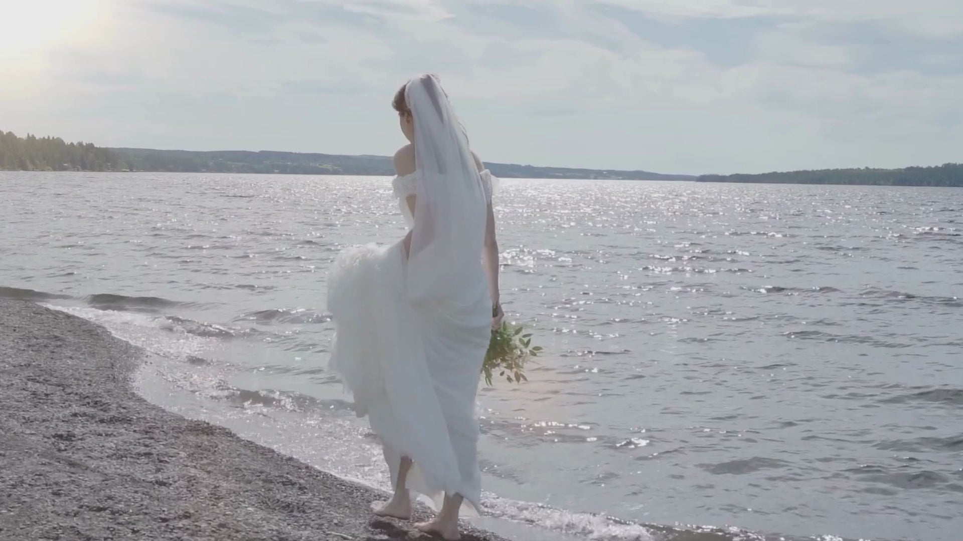 En brud kläd i vitt bröllopsklänning går på stranden med en blombukett i handen. 